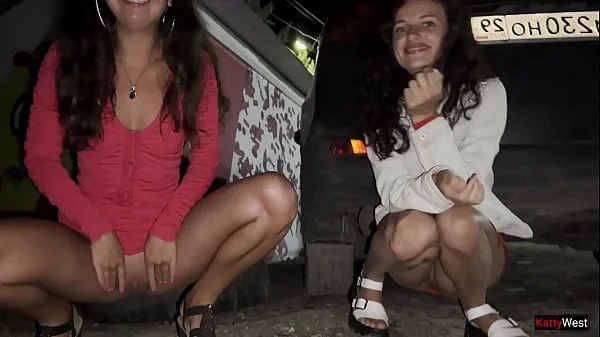 Two girlfriends pee together near a car in a public parking lot